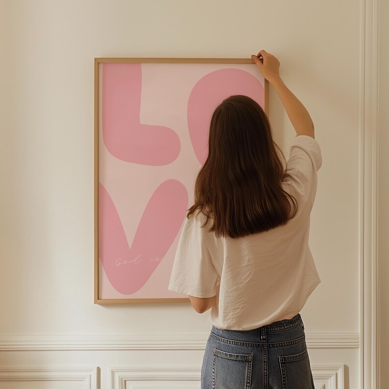May include: A framed pink and white abstract art print with the word "LOVE" in large, stylized letters. The print is hanging on a white wall.