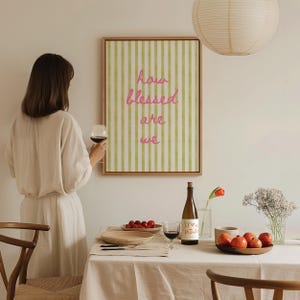 K&ouml;nnte beinhalten: Ein gerahmter Druck mit gr&uuml;nen und wei&szlig;en Streifen und dem Text "how blessed are we" in rosa Schrift. Eine Person h&auml;lt ein Glas Rotwein vor dem Druck. Eine Flasche Wein, ein Glas Rotwein und eine Schale rote &Auml;pfel stehen auf einem Tisch mit einer wei&szlig;en Tischdecke.