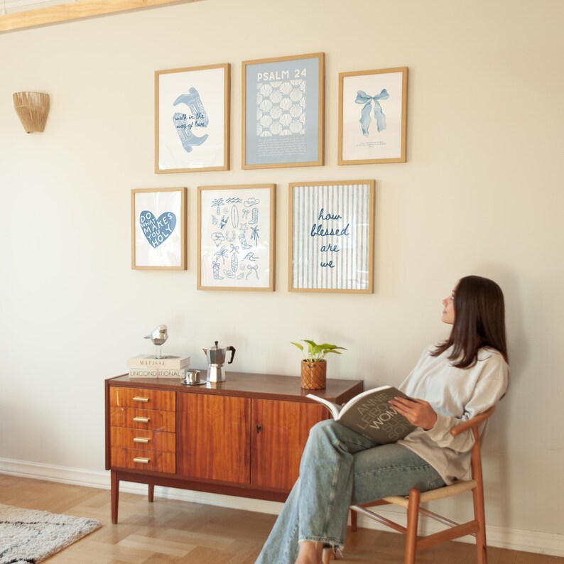May include: A display of framed art prints with blue and white designs, including text and illustrations, on a cream wall. Below, a wooden cabinet with drawers and doors. A person sits in a wooden chair, reading a book, creating a relaxed atmosphere.