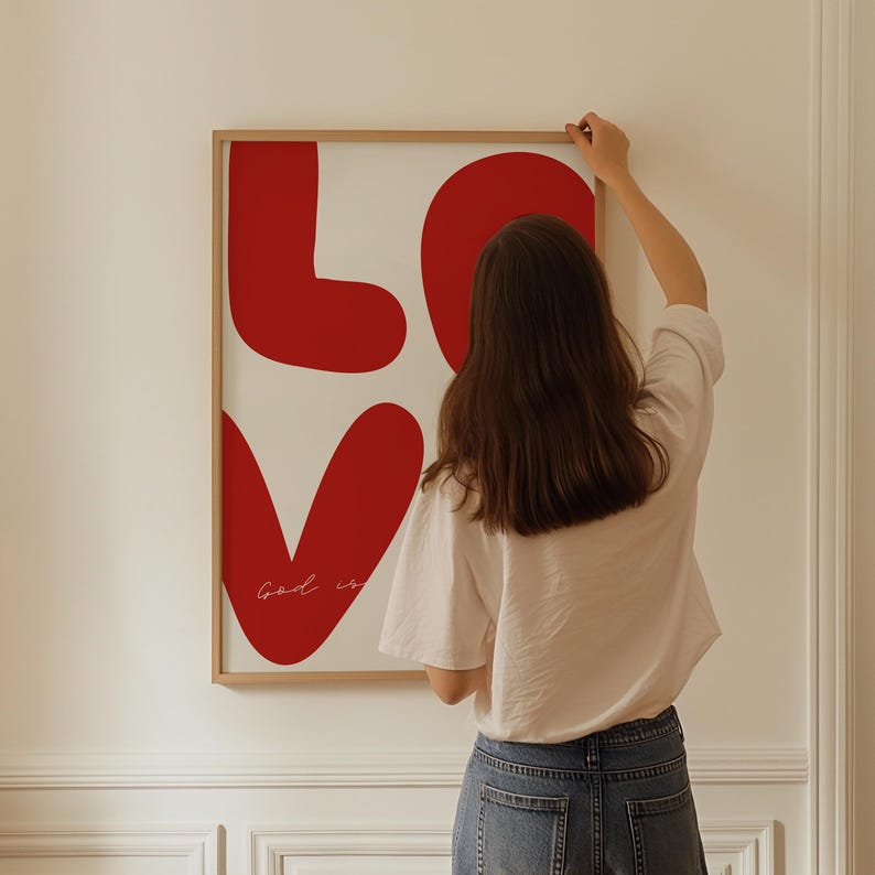 May include: A framed print with a white background and red stylized letters spelling "LOVE" with the text "God is" written in cursive inside the heart.