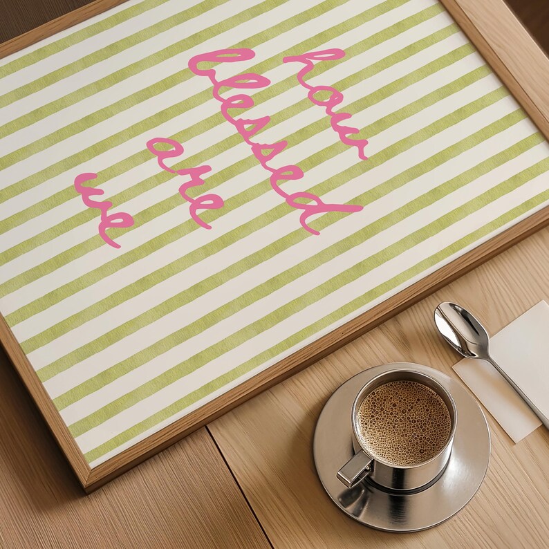 K&ouml;nnte beinhalten: Ein gerahmter Druck mit gr&uuml;nen und wei&szlig;en Streifen und dem Text "how blessed are we" in rosa kursiver Schrift. Der Druck liegt auf einem Holztisch mit einer Tasse Kaffee und einem L&ouml;ffel.