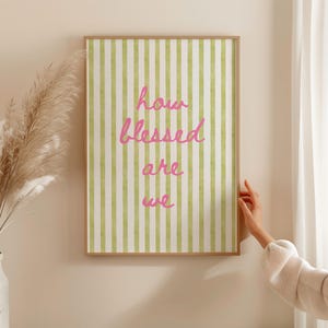 K&ouml;nnte beinhalten: Ein gerahmter Druck mit einem gr&uuml;n-wei&szlig; gestreiften Hintergrund und dem Text "how blessed are we" in rosa kursiver Schrift.