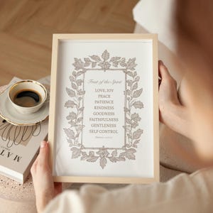 May include: A framed print with the text "Fruit of the Spirit" and a list of virtues in a decorative frame. The print is held by a person, with a cup of coffee and a book visible in the background.