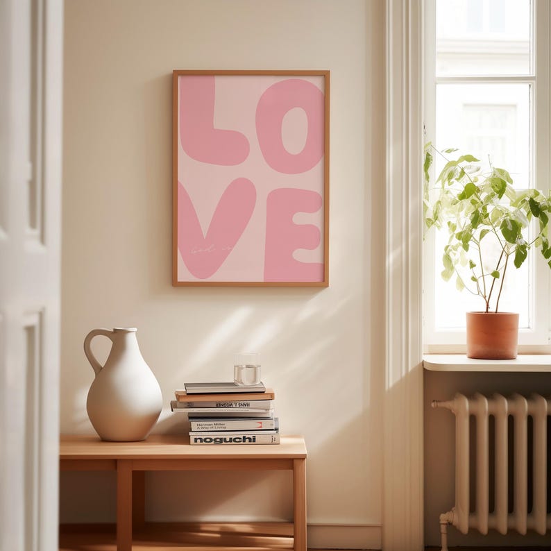 May include: A framed pink and white print with the word "LOVE" in large, bubble-like letters. The print is hanging on a white wall in a room with a wooden table and a potted plant.