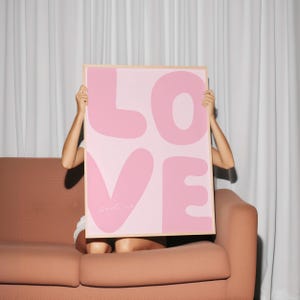 May include: A pink and white poster with the word "LOVE" in large, bubble-like letters. The poster is being held up by a person sitting on a brown couch.