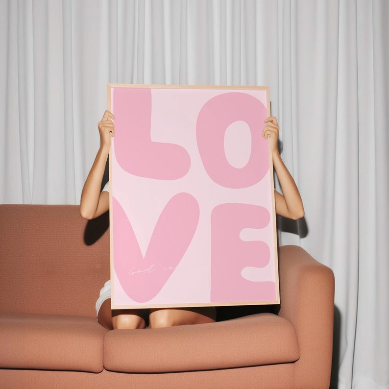 May include: A pink and white poster with the word "LOVE" in large, bubble-like letters. The poster is being held up by a person sitting on a brown couch.