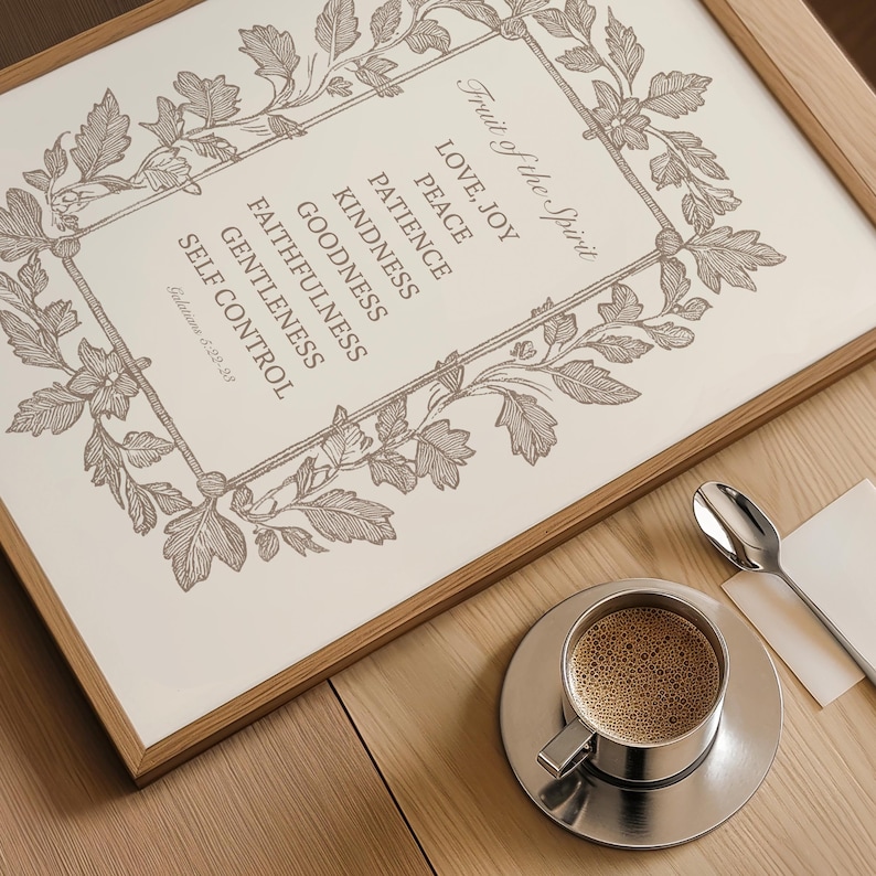 May include: A framed print with the text "Fruit of the Spirit" and related virtues, surrounded by a floral border. A silver coffee cup and saucer with coffee and a spoon are on a wooden table.