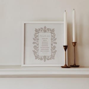 May include: A framed print with the text "Fruit of the Spirit" and a list of virtues. Two white candles stand in brass holders. The print is in a white frame, and the background is a neutral color.