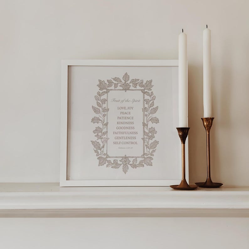 May include: A framed print with the text "Fruit of the Spirit" and a list of virtues. Two white candles stand in brass holders. The print is in a white frame, and the background is a neutral color.