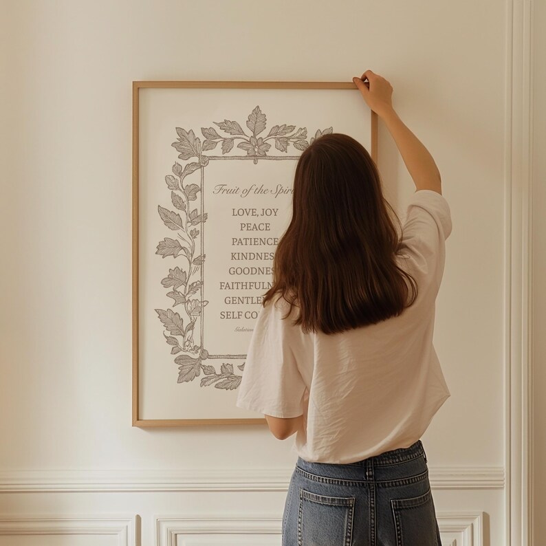 May include: A framed print with the text "Fruit of the Spirit" and a list of virtues in a decorative frame. The print is in a light color palette and is being hung on a white wall.