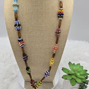 Puede incluir: Un collar de cuentas de colores con base de madera y borla. El collar presenta una variedad de colores, incluyendo rojo, azul, verde, amarillo y blanco. La borla está hecha de cuentas verdes y amarillas.