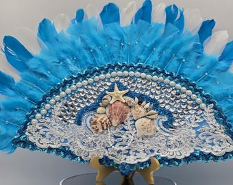 Peut inclure: Un éventail en plumes bleu et blanc avec un design en dentelle blanche et paillettes bleues. Le centre de l'éventail présente un motif de coquillages avec une étoile de mer et des perles.