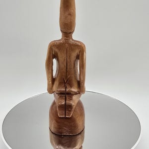 Pode incluir: Uma figura de madeira de uma pessoa sentada com as pernas cruzadas, com um chapéu pontudo. A figura é marrom e tem um acabamento liso. A figura está sentada em uma base redonda de cor prateada.