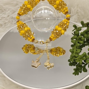 Puede incluir: Un brazalete de cuentas amarillas con un dije de corona dorada y un dije de mariposa dorada. El brazalete está sobre una superficie blanca con plantas verdes en el fondo.