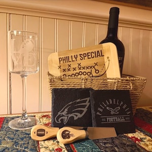 May include: A wine glass, cutting board, coasters, and cheese knives with a football theme. The cutting board reads "PHILLY SPECIAL" with a football play diagram. The coasters and knives feature the Philadelphia Eagles logo.