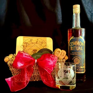 May include: A gift basket with a bottle of "Common Ritual" double barrel straight rye whiskey, a baseball-themed cutting board, coasters, a glass with a baseball diamond design, and a red bow. The basket is made of woven material.