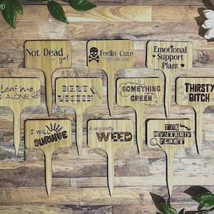 May include: A set of ten wooden plant markers with humorous sayings. The markers are made of natural wood and have a rustic look. The sayings include "Not Dead...yet", "Feelin' Cute", "Emotional Support Plant", "Leaf me Alone", "Next Victim", "Something Green", "I Will Survive", "A Very Pretty Weed", "The Mystery Plant", and "Thirsty Bitch".