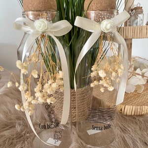 May include: Two clear glass bottles with cork stoppers and white ribbon bows. The bottles are decorated with black and white designs. One bottle says "Con bolle" and the other says "Senza bolle".