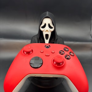May include: A red Xbox controller with white accents, featuring black buttons and a black directional pad. Behind the controller is a white Scream mask with a black hooded robe, creating a spooky theme.