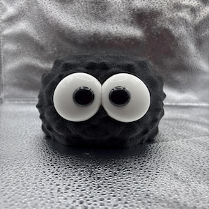 May include: A black, textured, cylindrical object featuring two large, round, white eyes with black pupils centered on the front. The surface of the object has a bumpy, uneven finish, and the overall shape is compact and rounded in appearance.