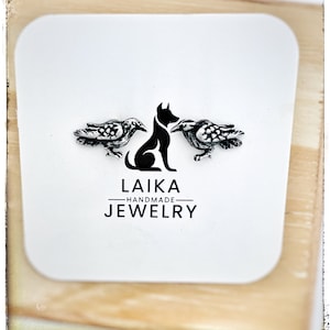 May include: A white card with the Laika Handmade Jewelry logo, featuring a black silhouette of a dog between two detailed raven figures. The text "LAIKA HANDMADE JEWELRY" is printed in black.