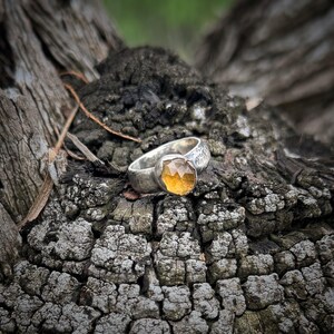 May include: A silver ring featuring a faceted, golden-yellow gemstone set in a bezel. The band of the ring has a textured, hammered finish, providing a distinct contrast to the smooth, geometric facets of the central stone.