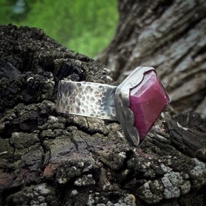 May include: A silver ring featuring a wide, hammered band with a textured finish and a single, faceted, deep pink gemstone set in a smooth, irregular metal bezel. The metal has a polished yet rustic appearance, highlighting the contrast between the hammered band and the smooth setting of the stone.