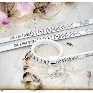 Op de afbeelding: Drie witte plastic ringmaatjes met zwarte markeringen. De maatjes zijn gemarkeerd met "SIZE & HALF SIZES" en hebben nummers van 1 tot 17. De maatjes liggen op een wit oppervlak met een bruin, pluizig voorwerp op de achtergrond.