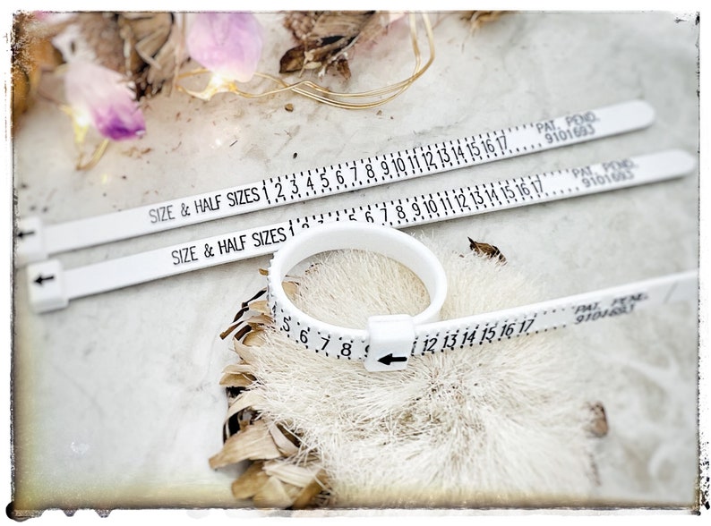 Op de afbeelding: Drie witte plastic ringmaatjes met zwarte markeringen. De maatjes zijn gemarkeerd met "SIZE & HALF SIZES" en hebben nummers van 1 tot 17. De maatjes liggen op een wit oppervlak met een bruin, pluizig voorwerp op de achtergrond.