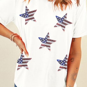 May include: White t-shirt with five star-shaped patches. Each star is decorated with a red, white, and blue American flag design.