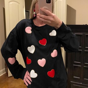 May include: Black long-sleeve sweatshirt adorned with various heart-shaped appliqués in pink, red, and white, each outlined with gold trim. The sweatshirt has a crew neck and a relaxed fit, suitable for casual wear.