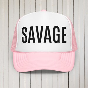 May include: A pink and white trucker hat with a black mesh back. The front of the hat has the word "SAVAGE" printed in black.