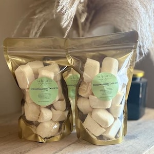 May include: Two clear, gold-lined pouches filled with white dishwasher tablets. Each pouch has a green label with the text "Dishwasher Tablets". The pouches are on a wooden surface, with dried ornamental grass in the background.