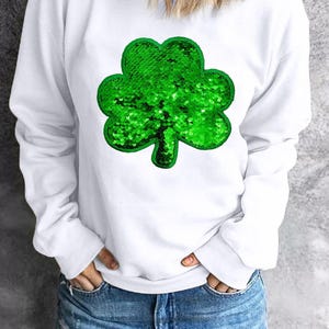 May include: White sweatshirt with a green sequin shamrock design. The sweatshirt has long sleeves and a crew neck.
