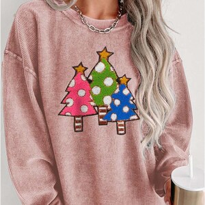 May include: A pink long-sleeved sweatshirt with a graphic of three Christmas trees with polka dots and stars. The trees are green, pink, and blue.