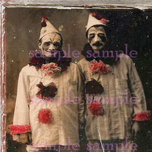 May include: Vintage black and white photograph of two clowns in costume. They wear white shirts, pants, and hats, with red and pink accents. Their faces are painted with dark makeup, and they stand side by side.