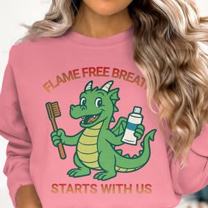 Op de afbeelding: Roze sweatshirt met een cartoon draak die een tandenborstel en tandpasta vasthoudt. De groene draak heeft gele accenten. De tekst "FLAME FREE BREATH STARTS WITH US" staat in oranje en geel boven en onder de draak.