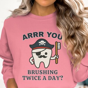 May include: Pink sweatshirt with a cartoon tooth wearing a pirate hat and eye patch, holding a toothbrush. The text on the shirt reads "ARRR YOU BRUSHING TWICE A DAY?"