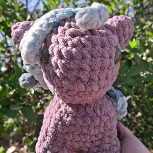 Puede incluir: Un juguete de peluche hecho a mano en tonos malva y gris. La criatura de ganchillo tiene grandes ojos verdes de botón y acentos esponjosos. El juguete es suave y texturizado, con un diseño caprichoso, ideal para una habitación infantil.