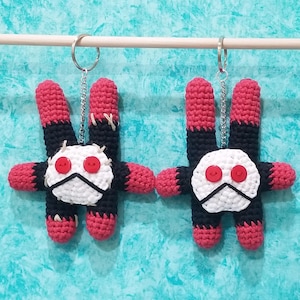 Pode incluir: Dois chaveiros de amigurumi crochetados em vermelho e preto com rostos brancos e olhos de botão vermelhos. Os chaveiros estão pendurados em correntes prateadas.