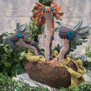 Puede incluir: Tres criaturas amigurumi de crochet que se asemejan a una planta con múltiples tallos y apéndices con forma de flor. Las criaturas son de color naranja, marrón y azul, con grandes ojos azules. Están sentadas en una maceta de crochet marrón.