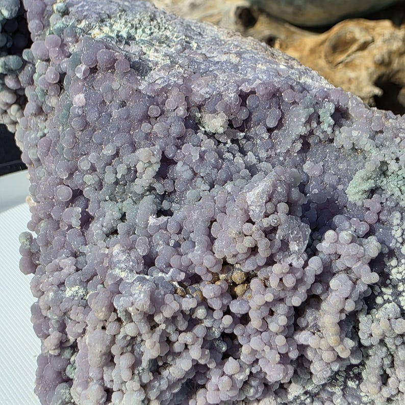 May include: A close-up of a large, rough-textured rock formation. The rock is primarily a soft purple color, with clusters of small, round, bead-like formations covering its surface. Hints of green and gray are visible.