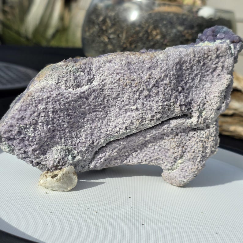 May include: A large, textured, lavender-colored rock formation with a rough, granular surface. The rock has a unique shape and is supported by a small, light-colored stone. The overall appearance suggests a natural mineral specimen.