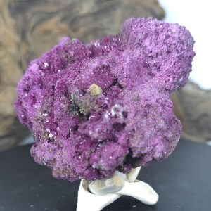May include: A vibrant purple mineral specimen with a rough, crystalline texture. The cluster features deep magenta hues with small, sparkling facets across its surface and a few scattered dark inclusions, supported by a white, multi-pronged stand.