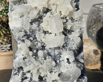 May include: A mineral specimen featuring a dark gray, textured base covered in clusters of small, translucent white crystalline formations. Several larger, distinct, clear cubic crystals are embedded among the smaller white mineral growths.
