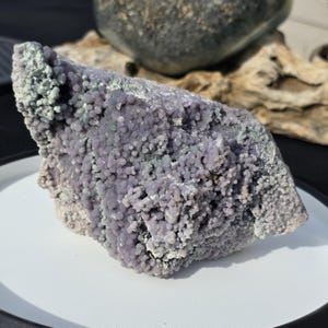 May include: A large, raw piece of grape agate, a unique mineral with a bubbly, botryoidal structure. The stone features a mix of lavender and pale green hues, with a rough, textured surface. The grape agate sits on a white surface.