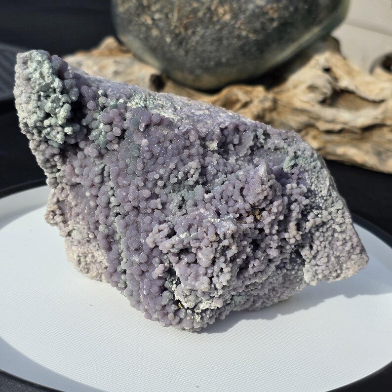 May include: A large, raw piece of grape agate, a unique mineral with a bubbly, botryoidal structure. The stone features a mix of lavender and pale green hues, with a rough, textured surface. The grape agate sits on a white surface.