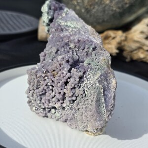May include: A large, irregular chunk of purple botryoidal chalcedony, a type of quartz. The surface is covered in small, rounded, grape-like formations. Some areas have a light green hue. The stone sits on a white plate.