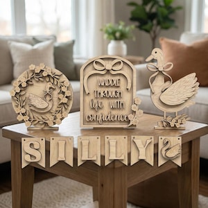 May include: Wooden swan ornaments, including a floral wreath, a sign reading "Waddle Through Life With Confidence", and a banner spelling "SILLY". The swans are light brown and displayed on a wooden table.