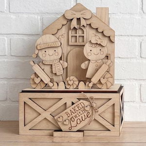 Könnte beinhalten: Eine dekorative Box aus hellem Holz mit einer lasergeschnittenen Lebkuchenhaus-Szene und zwei Lebkuchenfiguren, die Nudelhölzer halten. Ein mit Bindfaden befestigtes Holzschild auf der Vorderseite trägt die Gravur Baked with Love.